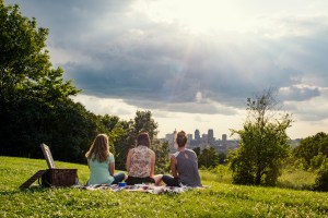 Three women sitting in a park, cityscape in the background.