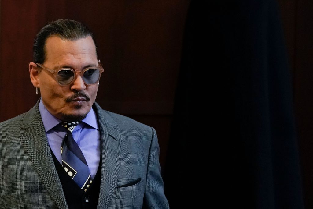 Johnny Depp, wearing sunglasses, a grey suit, a dark vest and a patterned blue tie, walks into the left side of the frame.