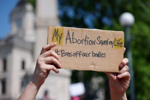 A white person's hands are shown holding a handwritten cardboard sign reading "My abortion saved my life, #bans off our bodies."