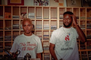 Vonne Parks and Andre Gainey of They Hate Change stand against a shelf filled with CD cases, wearing white shirts.