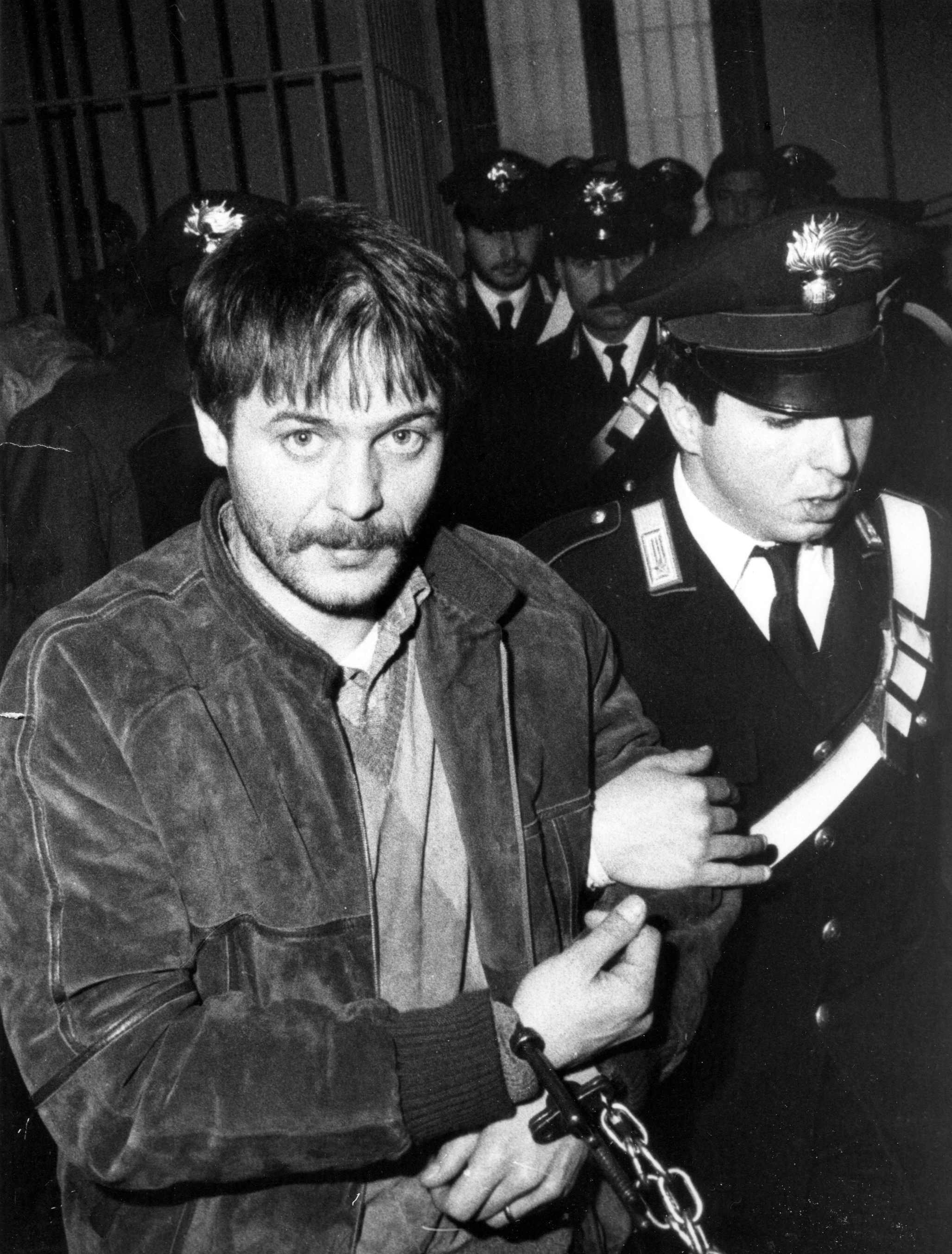 Renato Vallanzasca – Man with a 70s moustache, wearing a leather bomber jacket and a sweater vest, being carried away by policemen with his hands cuffed.