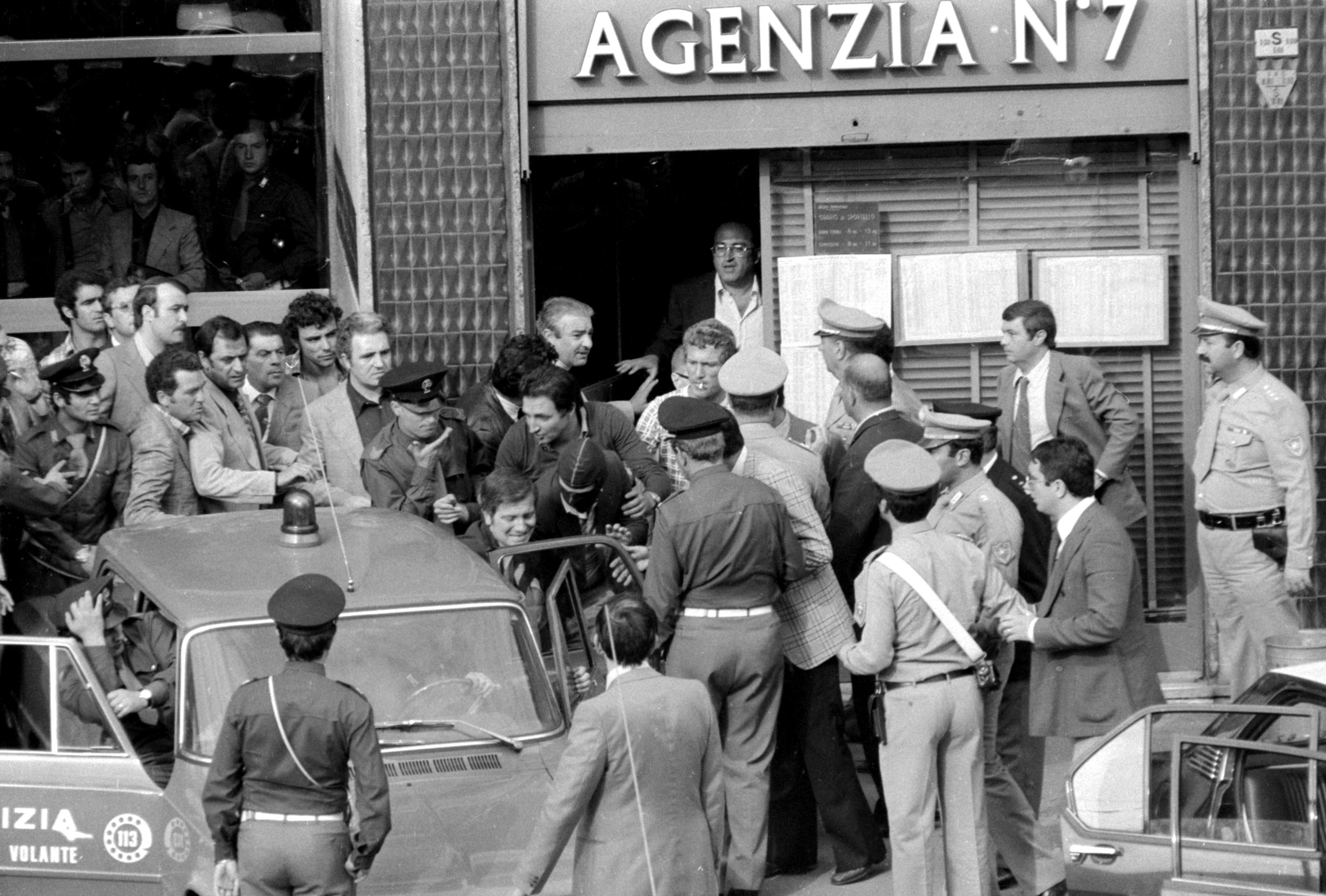 Robbery Piazza Insubria – black and white picture of a group of men pushing someone with a hood over their head into a police car.