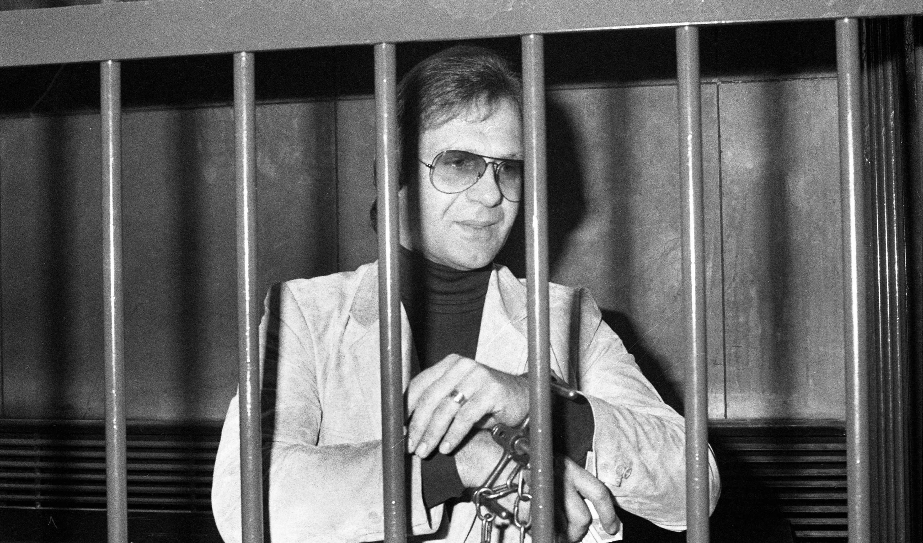 Francis Turatello – black and white photo of a man wearing a jacket, a turtleneck and shaded aviator sunglasses behind bars.