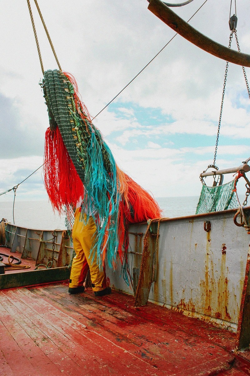 Ostend, fishing, photography - Photograph of the back of a fisherman wearing yellow dungarees and holding a colourful fishing net.