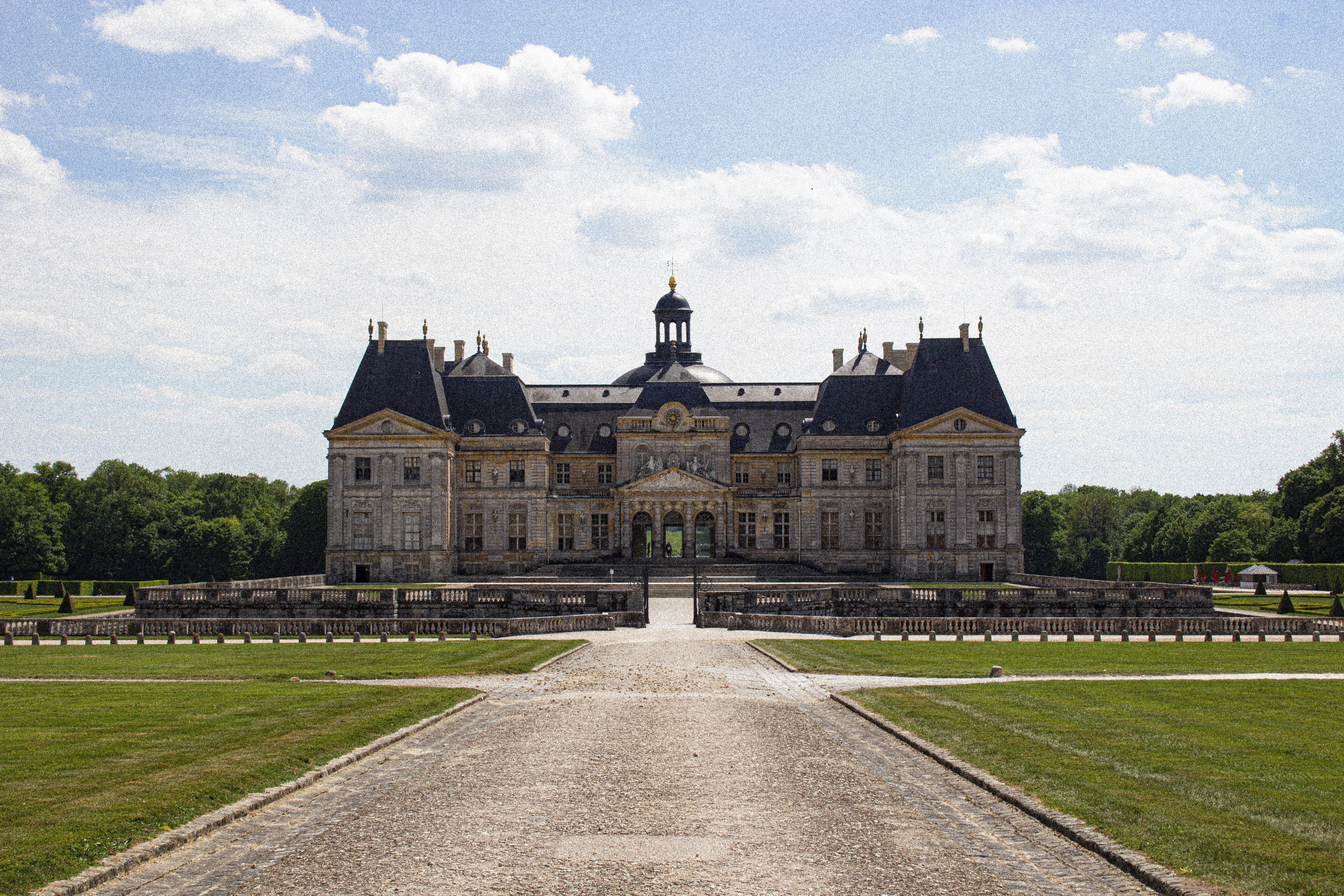 Château de Vaux-le-Vicomte – photo of a large castle in baroque style, made out of stone with gold accents and an anthracite roof, laying the middle of a large garden.