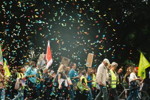 Ein Gruppe Menschen schreitet mit Warnwesten und Fahnen durch Konfettiregen. Es sind Klinikangestellte in Köln, die streiken.