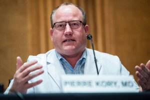 Photo shows Pierre Kory in a lab coat in front of a microphone. his hands are raised as he talks.