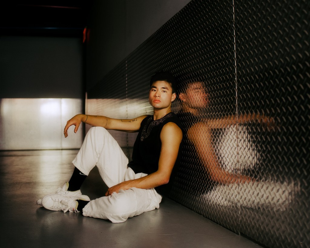 Chella Man leans against a wall, sitting with his arm on his knee.