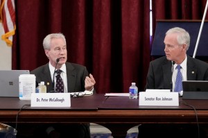 Photo shows Peter McCullough, left, speaking as Ron Johnson, right, looks on. Both men are white and have white hair and are wearing suits, sitting in front of microphones and name plates.