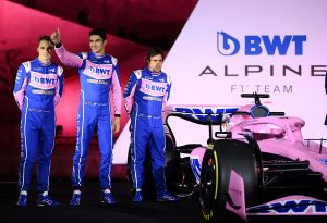 Oscar Piastri, Esteban Ocon, and Fernano Alonso stand next to the pink Aston Martin F1 car. Alonso has a sour expression.