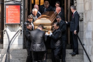 A gold casket is shown being guided down the stone steps of a church by numerous pallbearers wearing black suits and white gloves.