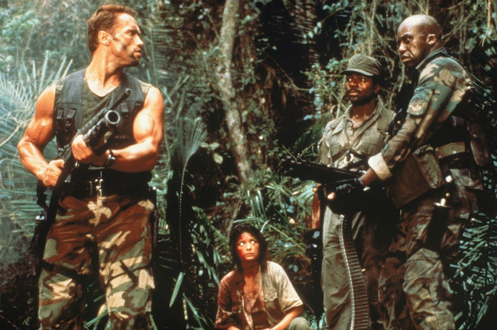 Still shot from Predator - Dutch, holding a large gun, looks over to Mac and Dillon as Anna sits on the Jungle floor between them.