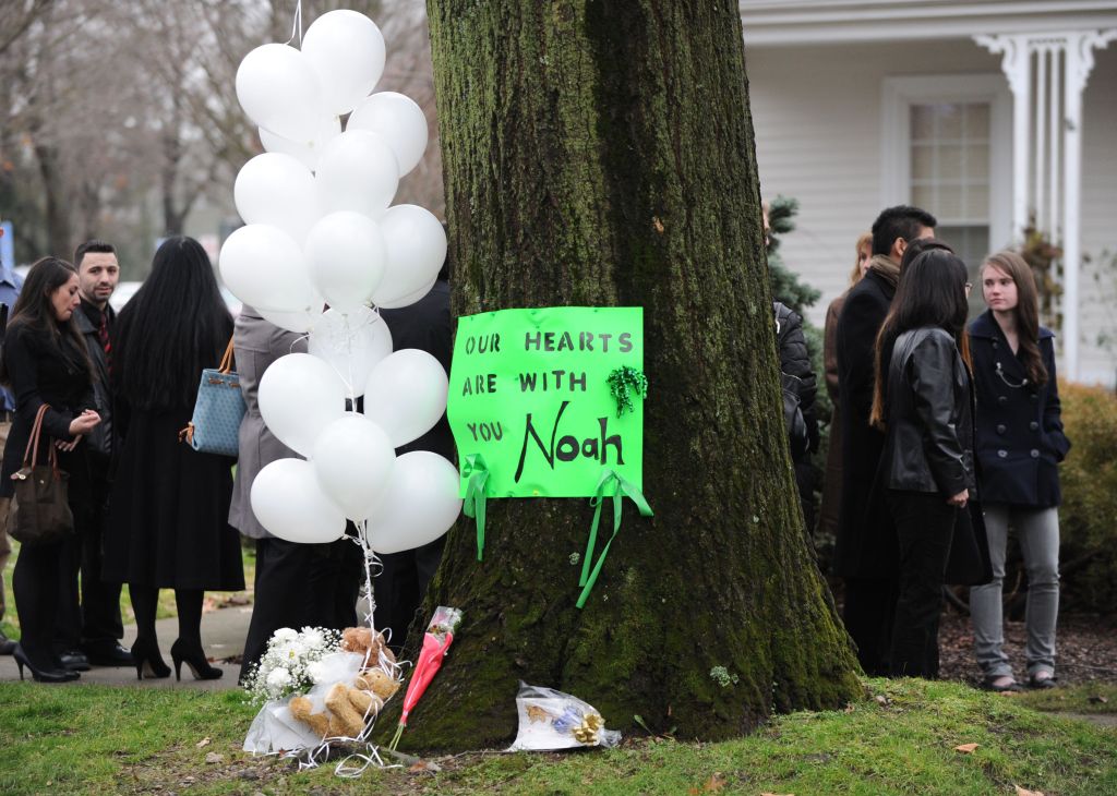 Photo shows a tree with balloons and a sign in front of it that reads "Our hearts are with you, Noah." There are a crowd of people dressed in dark colors on either side of the tree, just behind it.