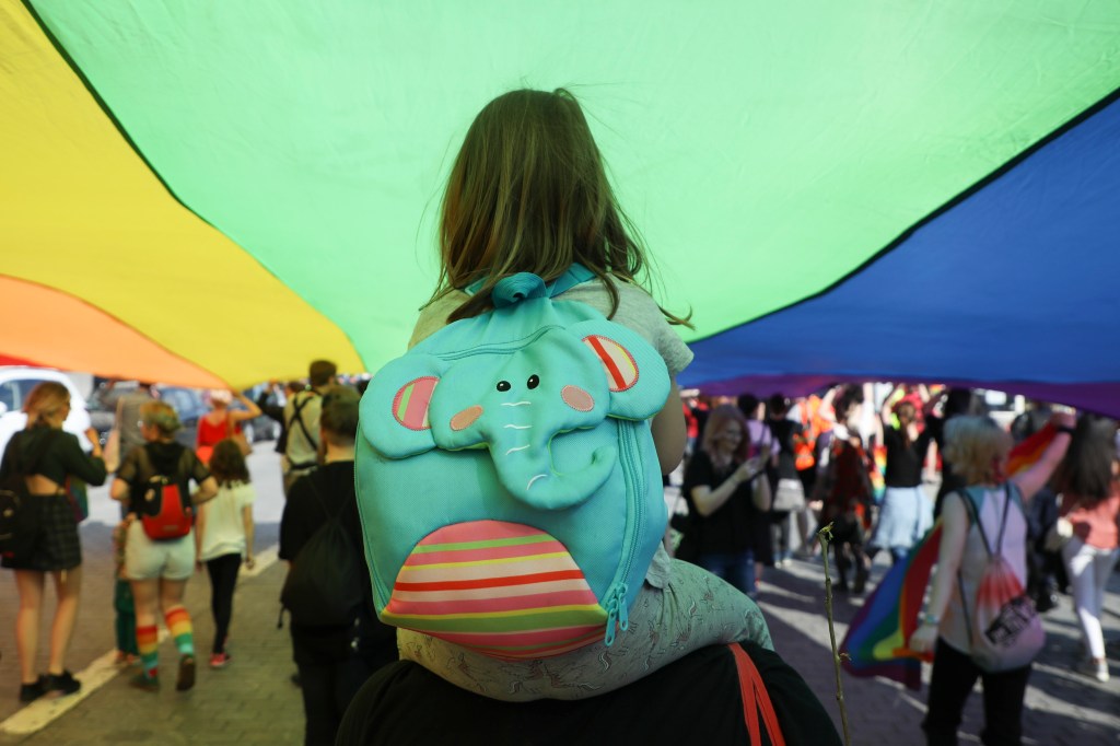 A long-haired child wearing an elephant backpack sits on the shoulders of a parent under a rainbow umbrella at a LGBTQ Pride march in Krakow, Poland