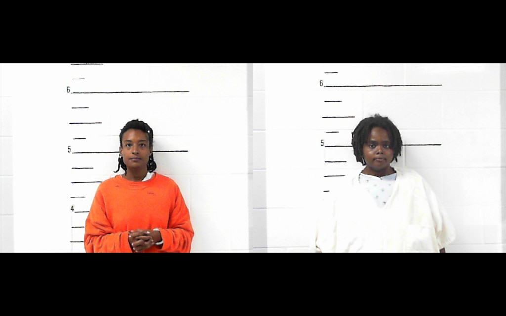 Photo shows two women's booking photos; both stand in front of height charts. the woman on the left is Black and has an orange sweatshirt. The woman on the right is also Black and has a white draped gown on overa hospital garment.