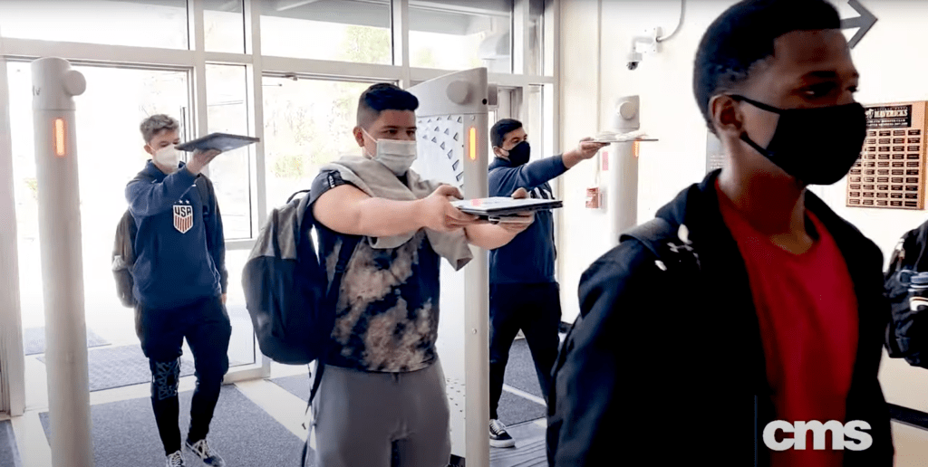 Students going through a weapons scanner made by Evolv Technology while holding laptops and notebooks in front of them.