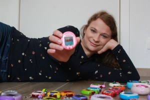 Tamagotchi – young woman lying on the ground surrounded by tamagotchis in different colour, showing one of them to the camera