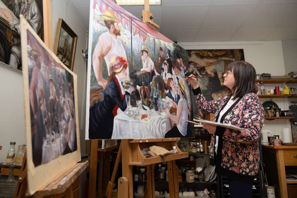 Andréa Dlouha, reproduction artist –middle-aged woman standing in front of a large painting propped on an easel, carefully adding details with a brush.