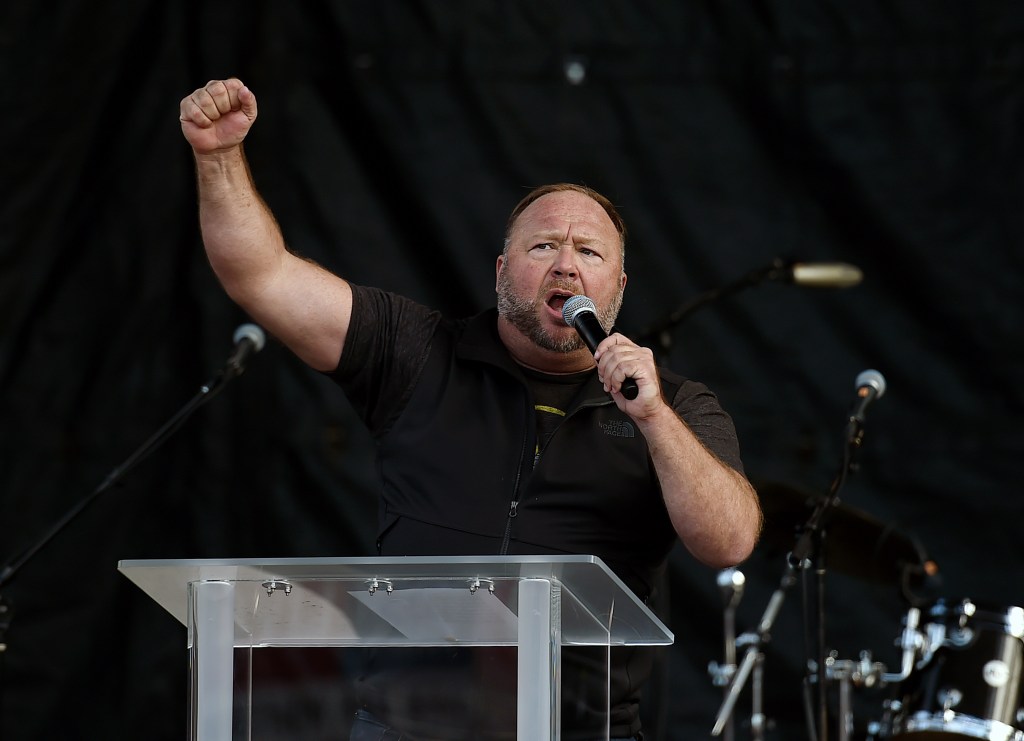 Alex Jones stands behind a podium holding a microphone with one fist raised in the air.