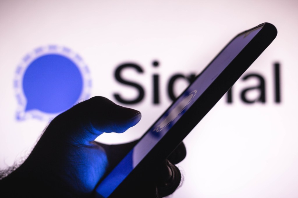 In this photo illustration the Signal logo seen in the background of a silhouette hand holding a mobile phone.