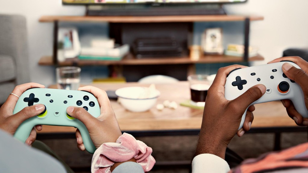 Two people hold stadia controllers while sitting in front of a small coffee table.