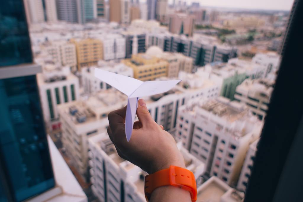 Photo of a hand wearing an orange watch throwing a paper plane out of a window. Background: highrises.