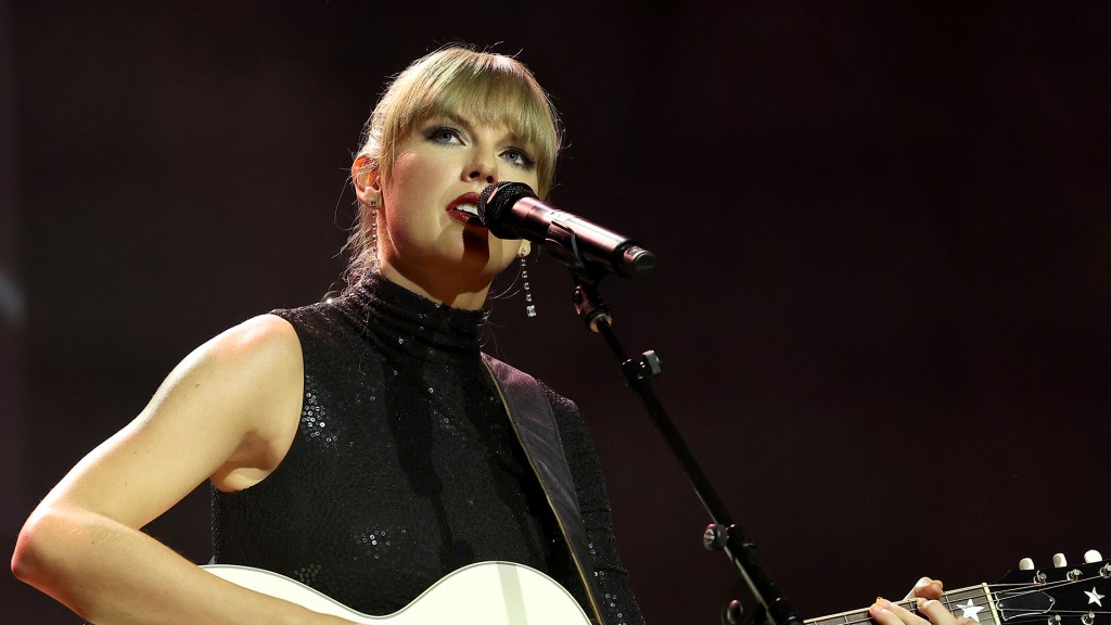 Taylor Swift plays the guitar onstage as she sings into a microphone