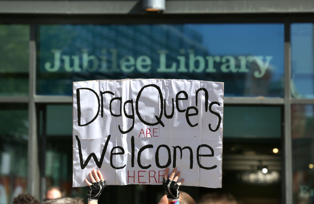 a sign that reads "Drag Queens are Welcome Here" is held in front of a library entrance