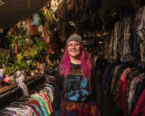 GRIN shop manager, Freya McGreevy, standing in front of rails of vintage clothes. She has pink hair, glasses and a beanie hat.