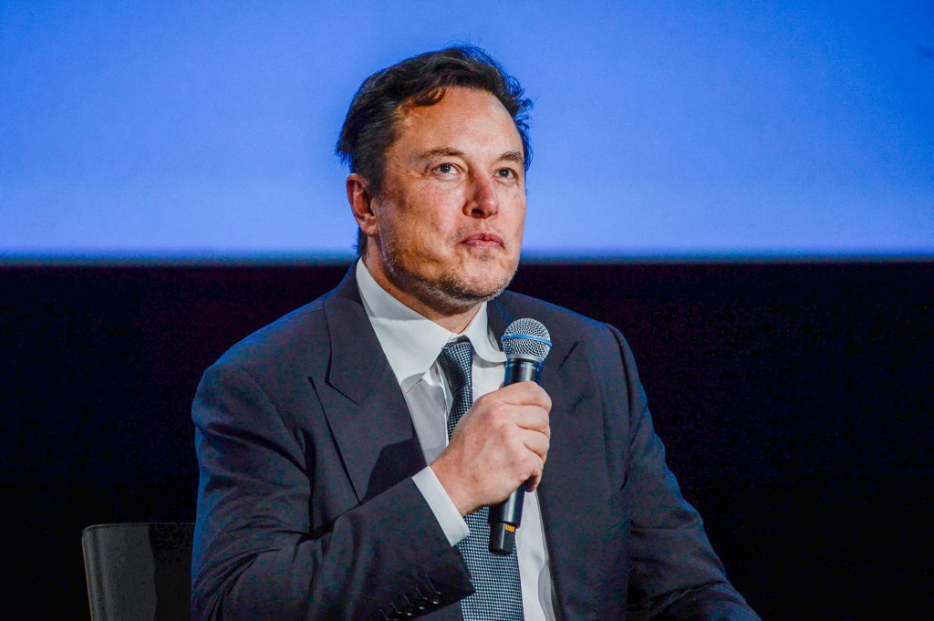 Elon Musk looking sweaty in a suit and tie holding a microphone seated on a stage.