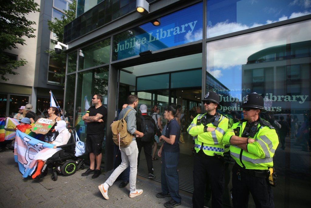 Two police officers stand next to a crowd of people waving LGBTQ banners outside a library.