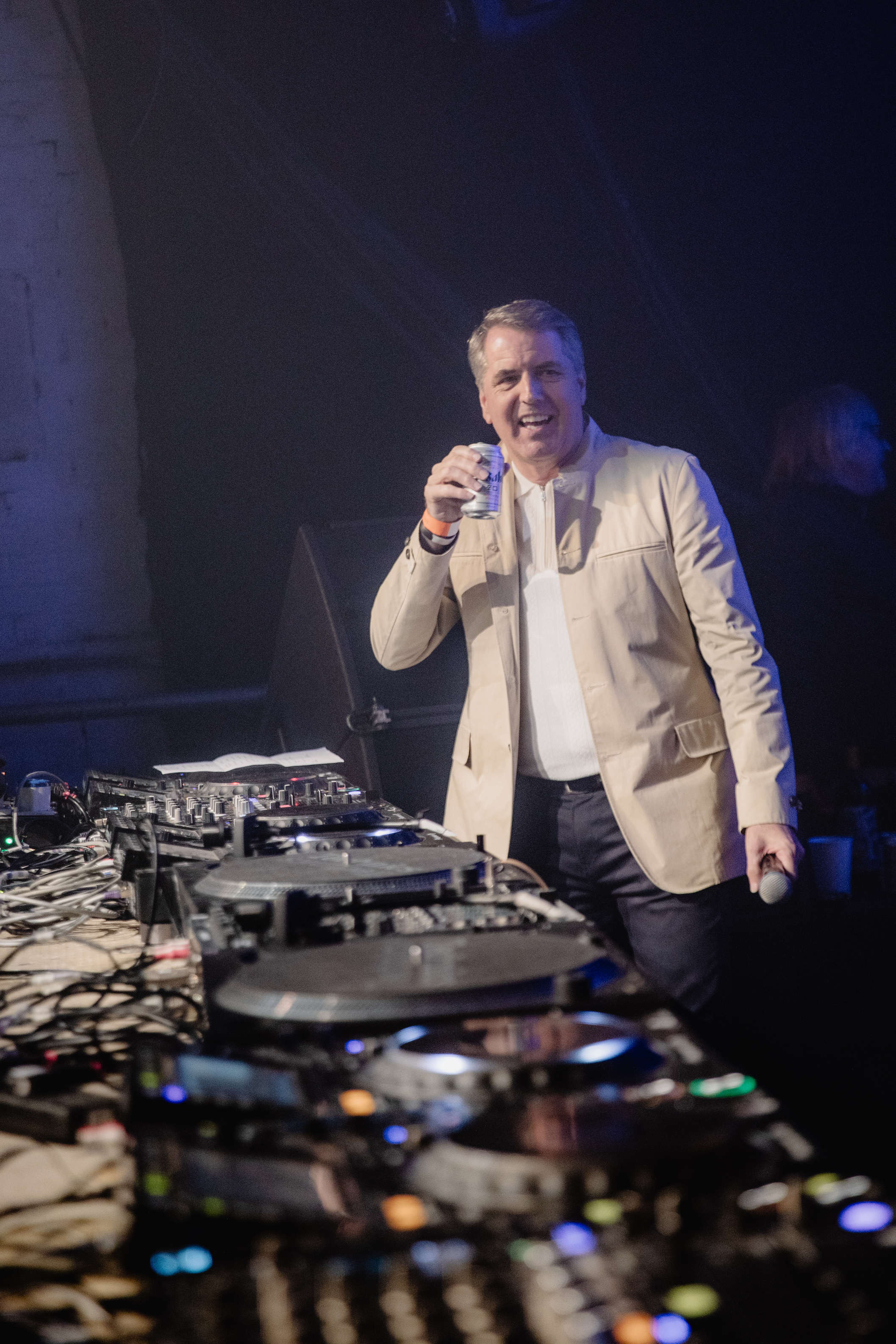 Steve Rotheram smiles behind the dj decks, wearing a cream jacket, white top and jeans, holding a can of beer.