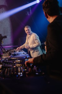 Steve Rotheram smiles behind the dj decks, wearing a cream jacket, white top and jeans, holding a can of beer.