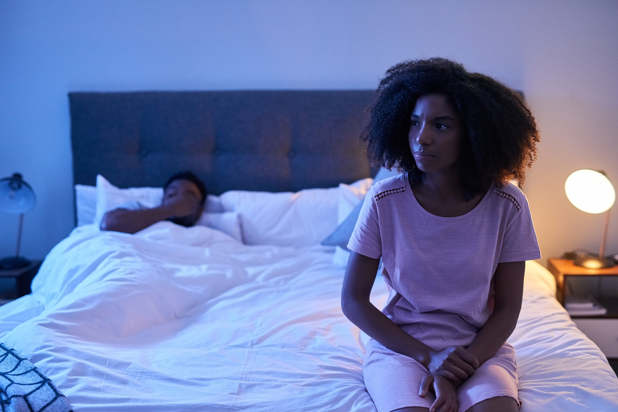 A woman sat on edge of bed looking sad while her boyfriend lies in bed looking stressed.