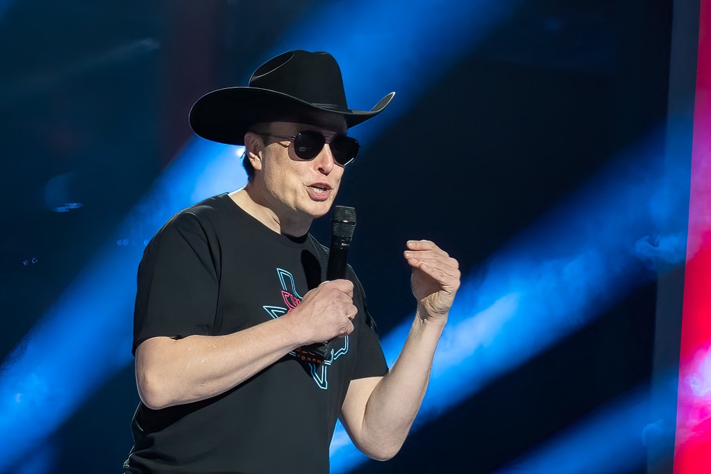 Elon Musk speaks onstage with a black cowboy hat on and sunglasses. He's holding a microphone.