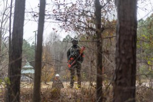 A police officer wearing black riot gear and armed with a rifle stands just outside the tree line of the Atlanta Forest.