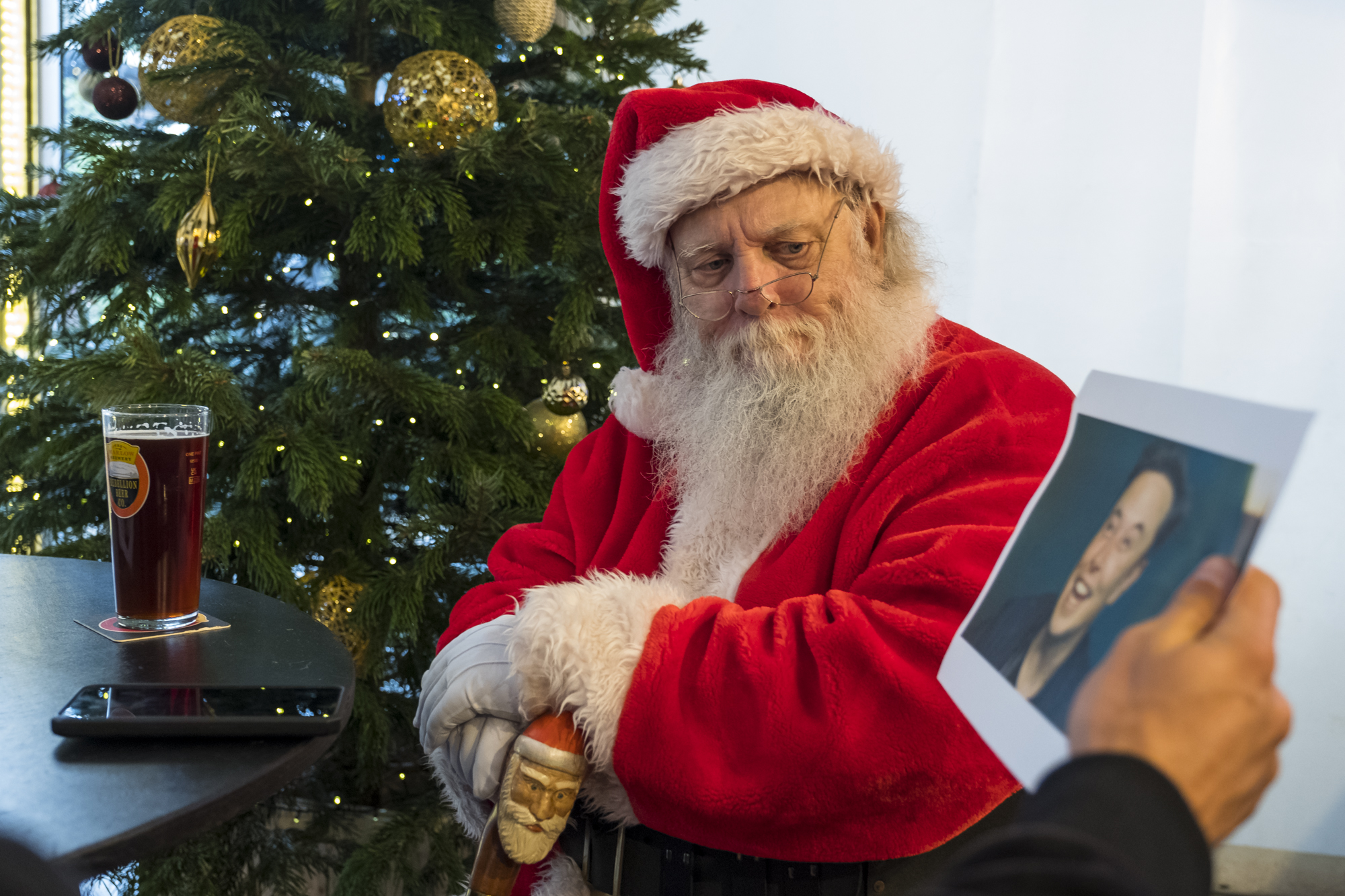 A man dressed as Santa in the red outfit sat on a table with a pint of beer looking at a photo of Elon Musk.