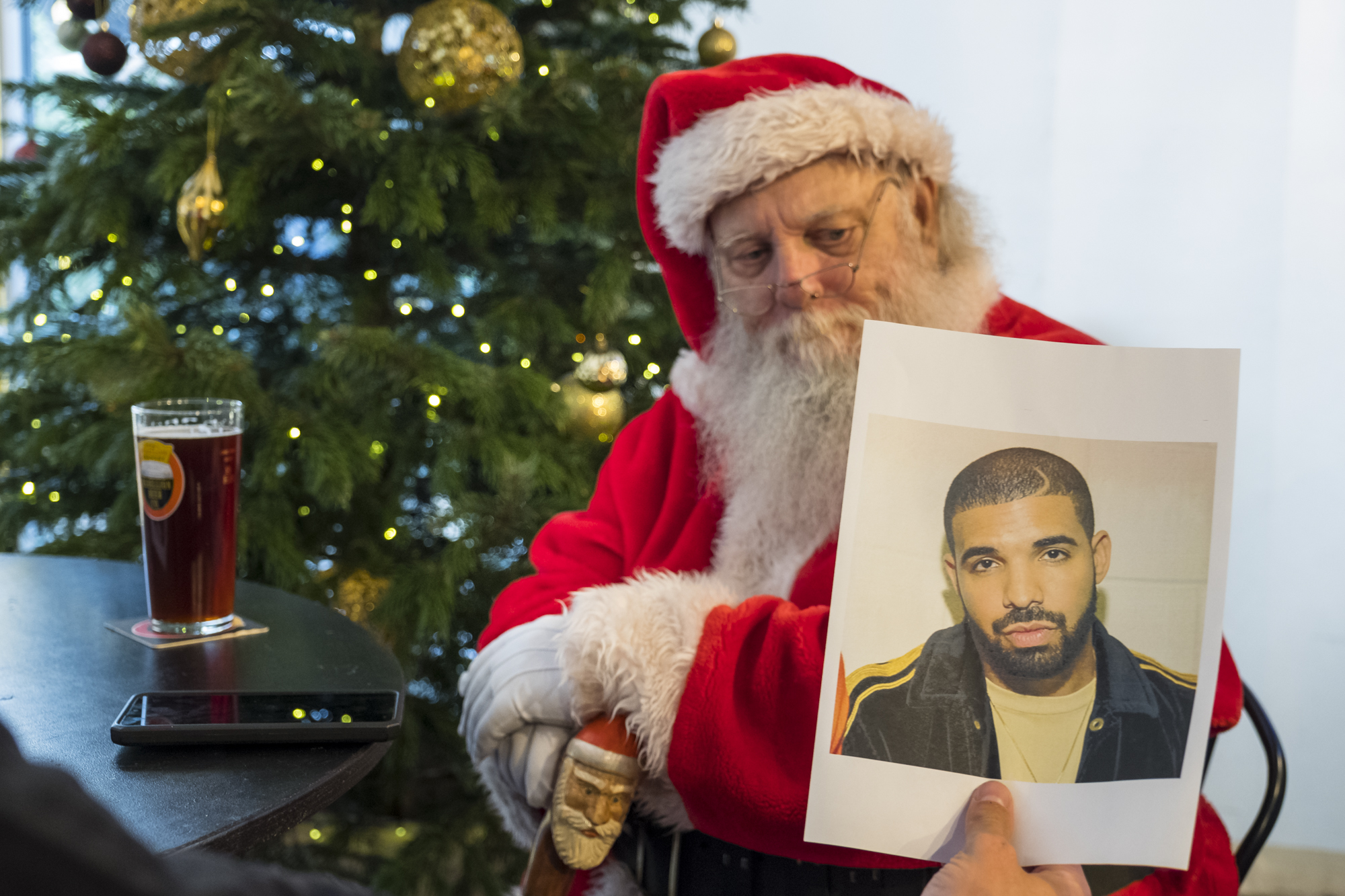 A man dressed as Santa in the red outfit sat on a table with a pint of beer being passed at a photo of Drake.