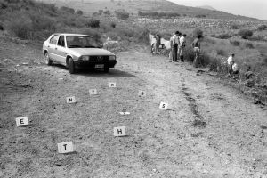 Fabio Sgroi, second mafia war – group of people on a hill with a car parked next to them. On the ground, there are numbered police signposts to mark the crime scene.