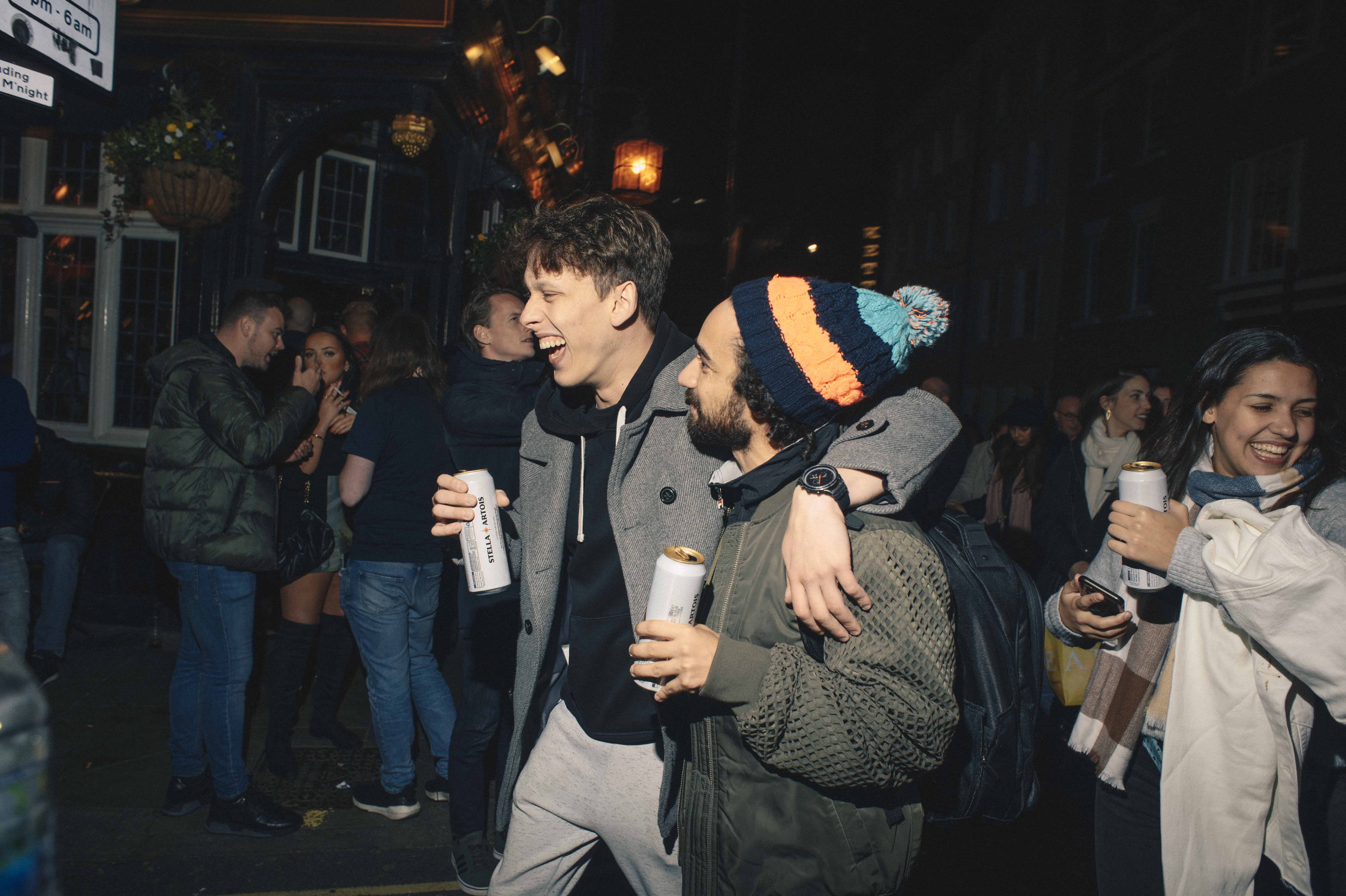 Two men walking on a busy street with beer cans in hand, they are embracing each others shoulders.