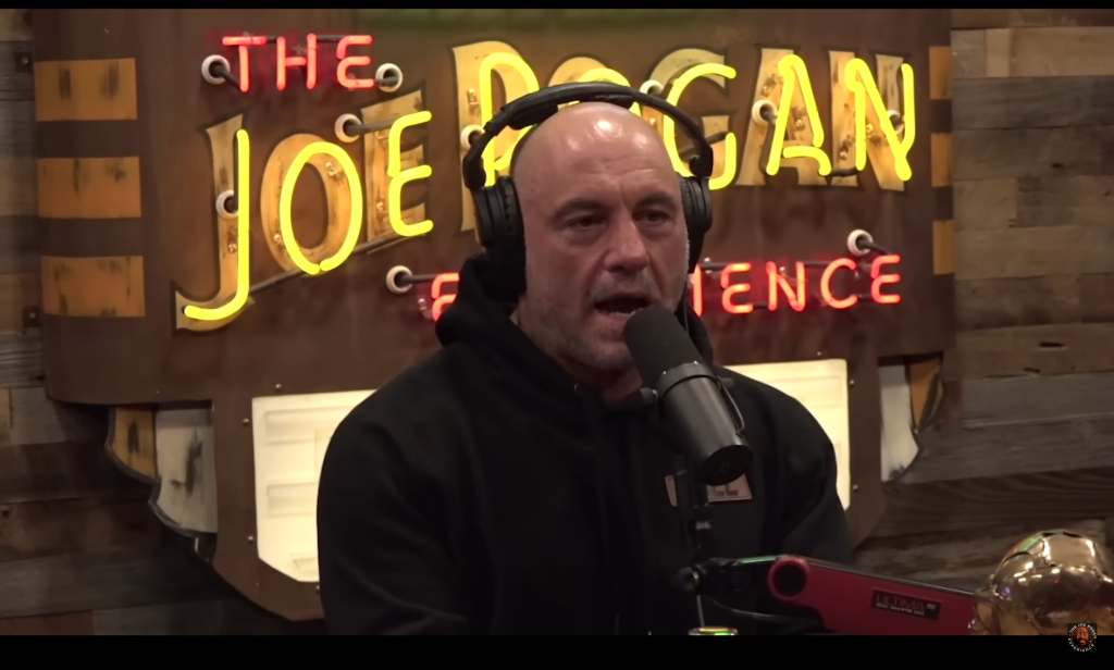 Joe Rogan wears a black sweatshirt and sits in front of a microphone.