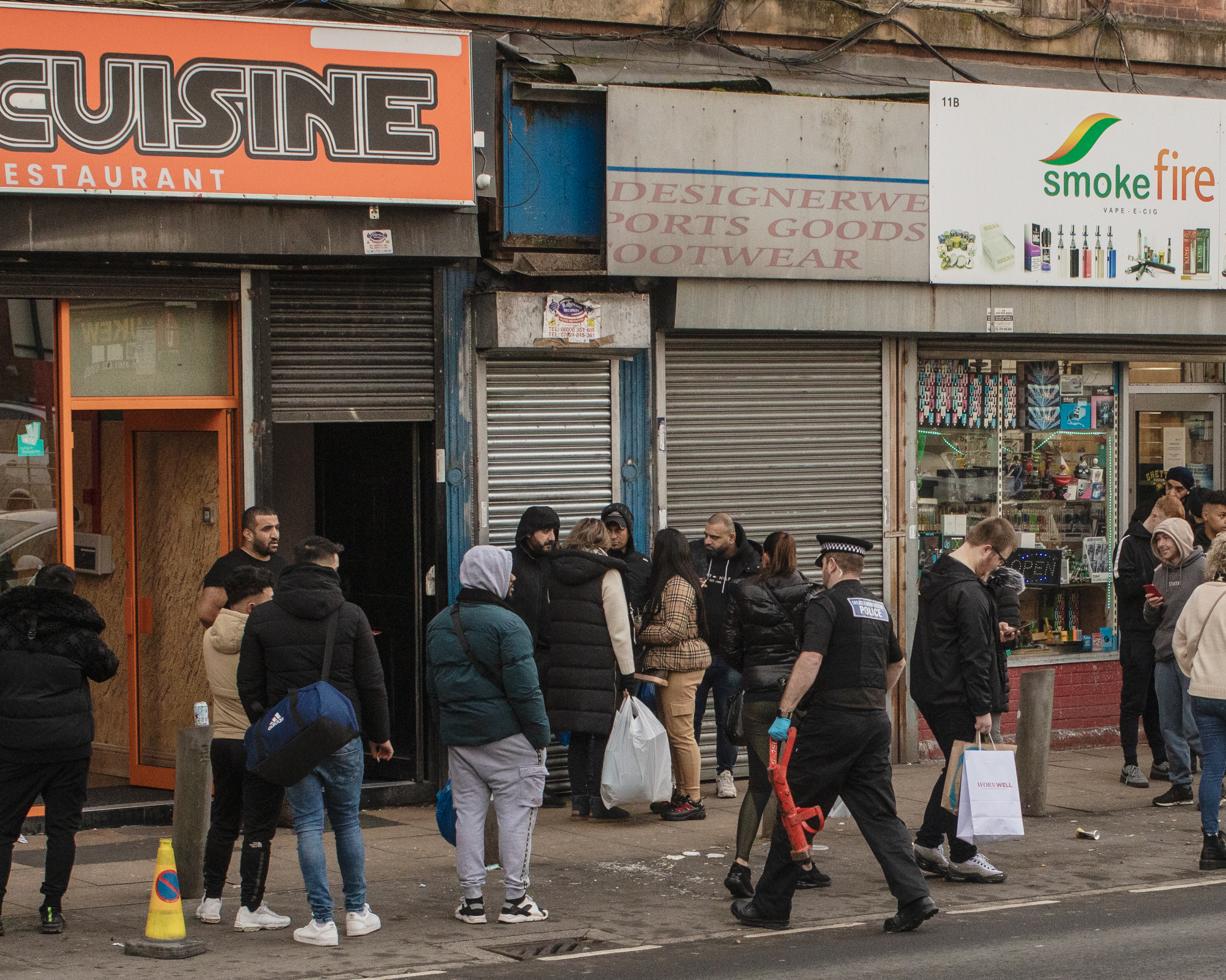 People convene outside of a shuttered store next to a restaurant, a police officer is walking up in the foreground.