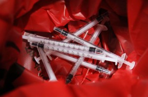 Image shows a pile of used vaccine syringes in a red medical waste bag.