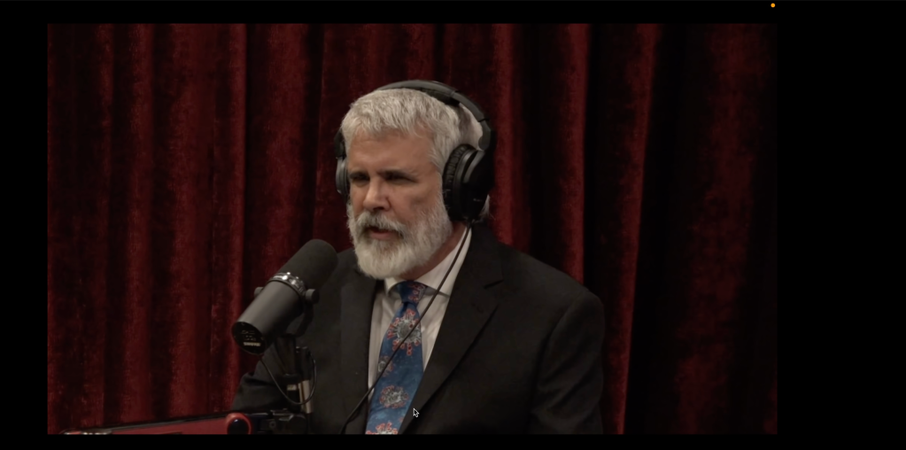 Robert Malone sits at a microphone. He has a white beard and a patterned tie with a lot of blue in it.