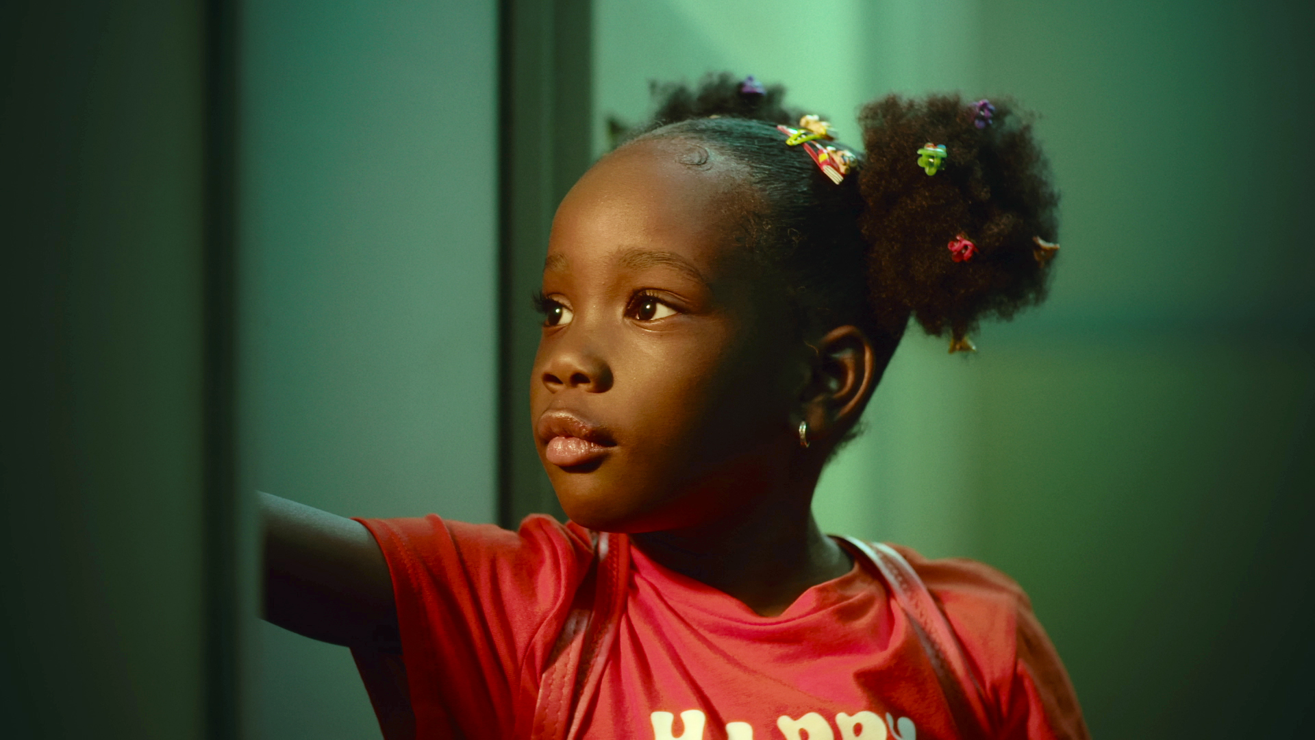 Elisabetta Agyeiwaa – image of a young girl with afro puffs and lots of hair clips, standing in a dimly lit room and looking off camera