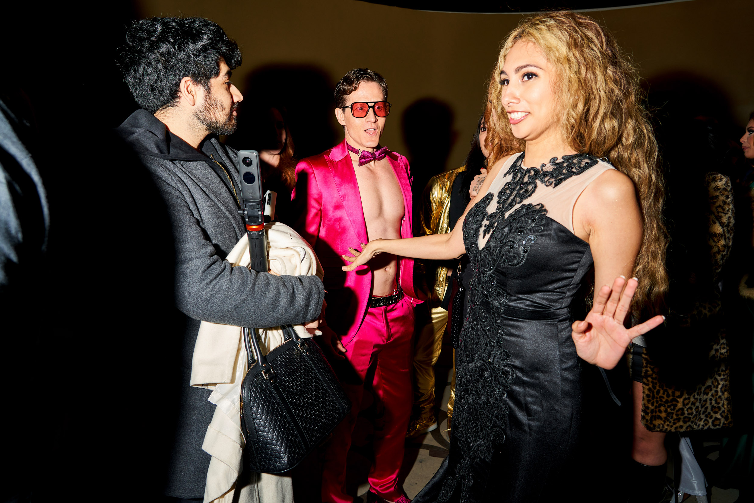 A shirtless man in a pink suit and a bowtie looks at a woman in the foreground with a surprised face.