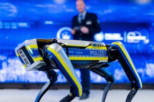 A quadrupedal robot with blue and yellow police markings stands in front of a blue background.