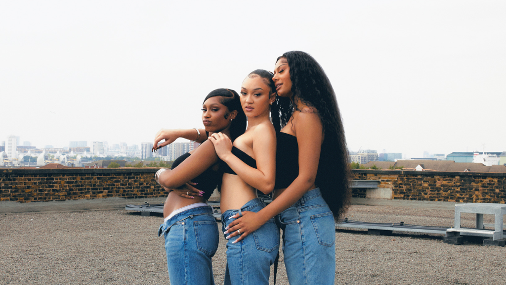 Three Black members of girl group Flo stand in a lon a roof in jeans and black tube tops.