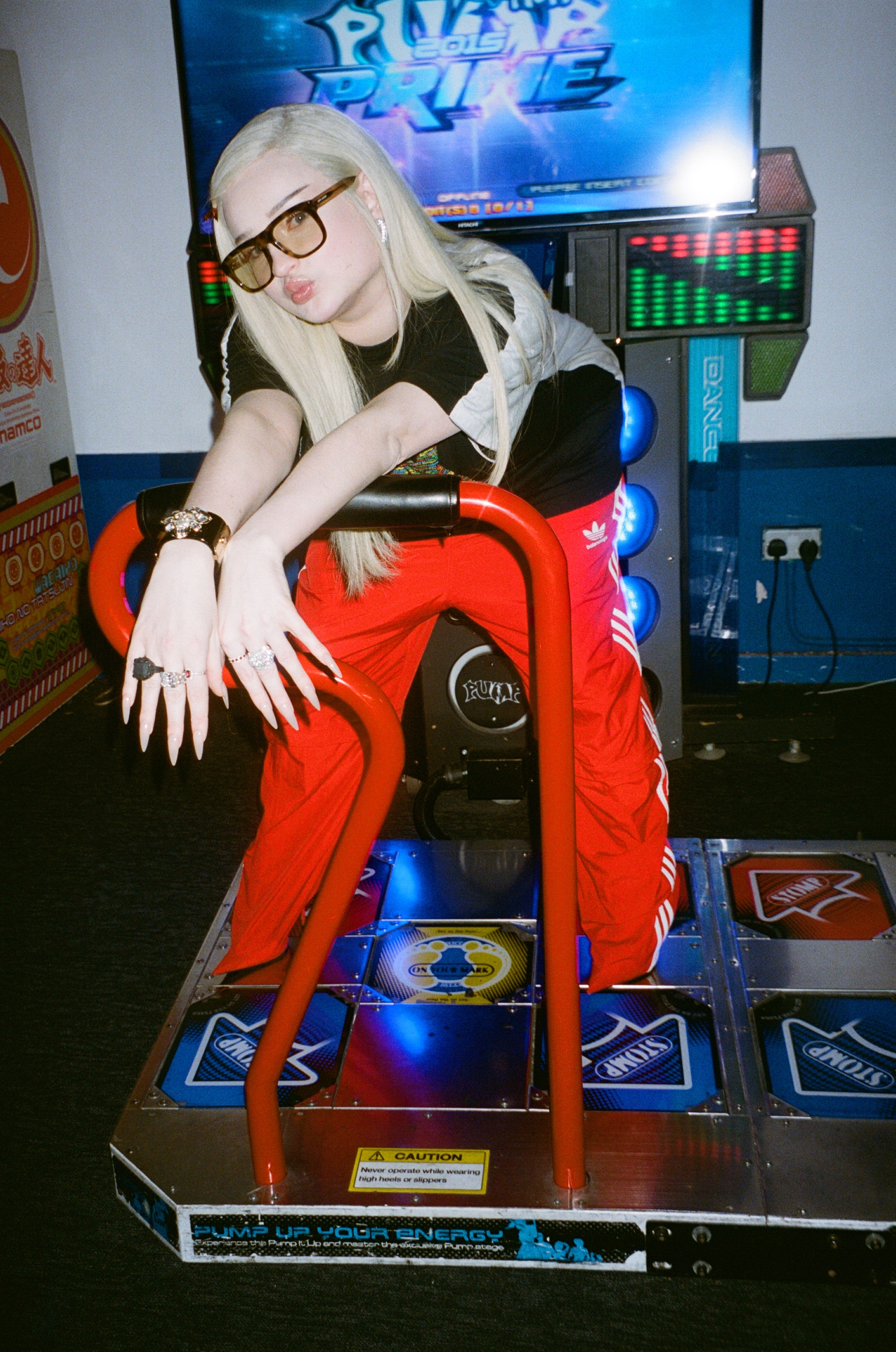 Kim Petras leans over a Dance Dance Revolution arcade machine with a pouty face sporting long nails.