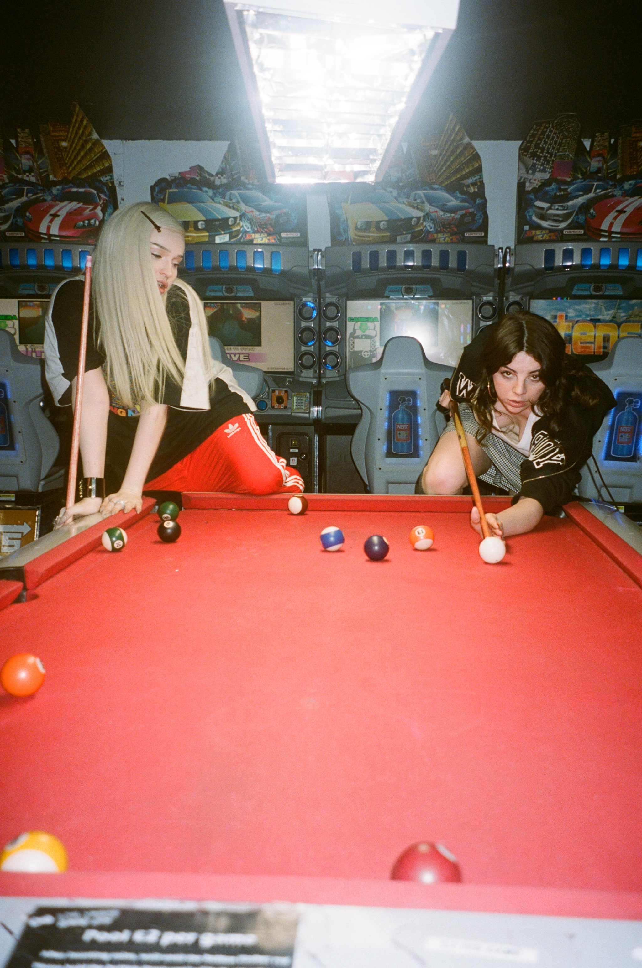 Kim Petras leans over a pool table with Shon while Shon lines up a shot and looks at the camera.